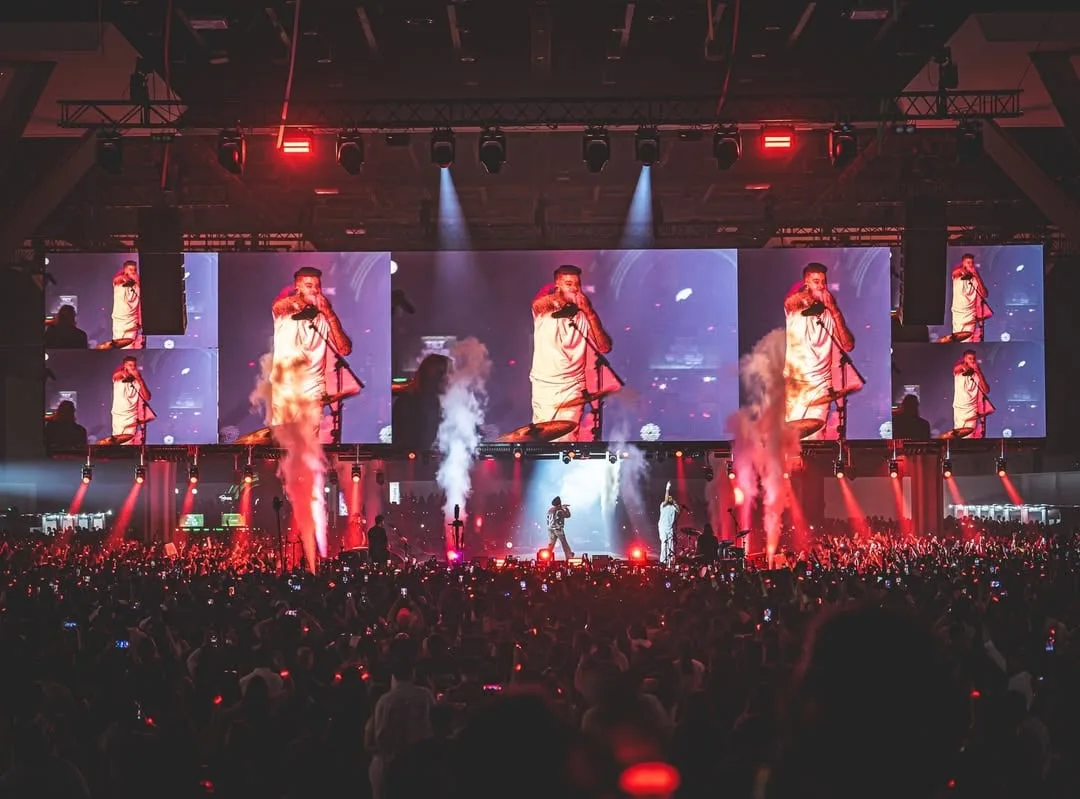 Indoor arena with stacked screen bays showing the artist, red and violet light through smoke
