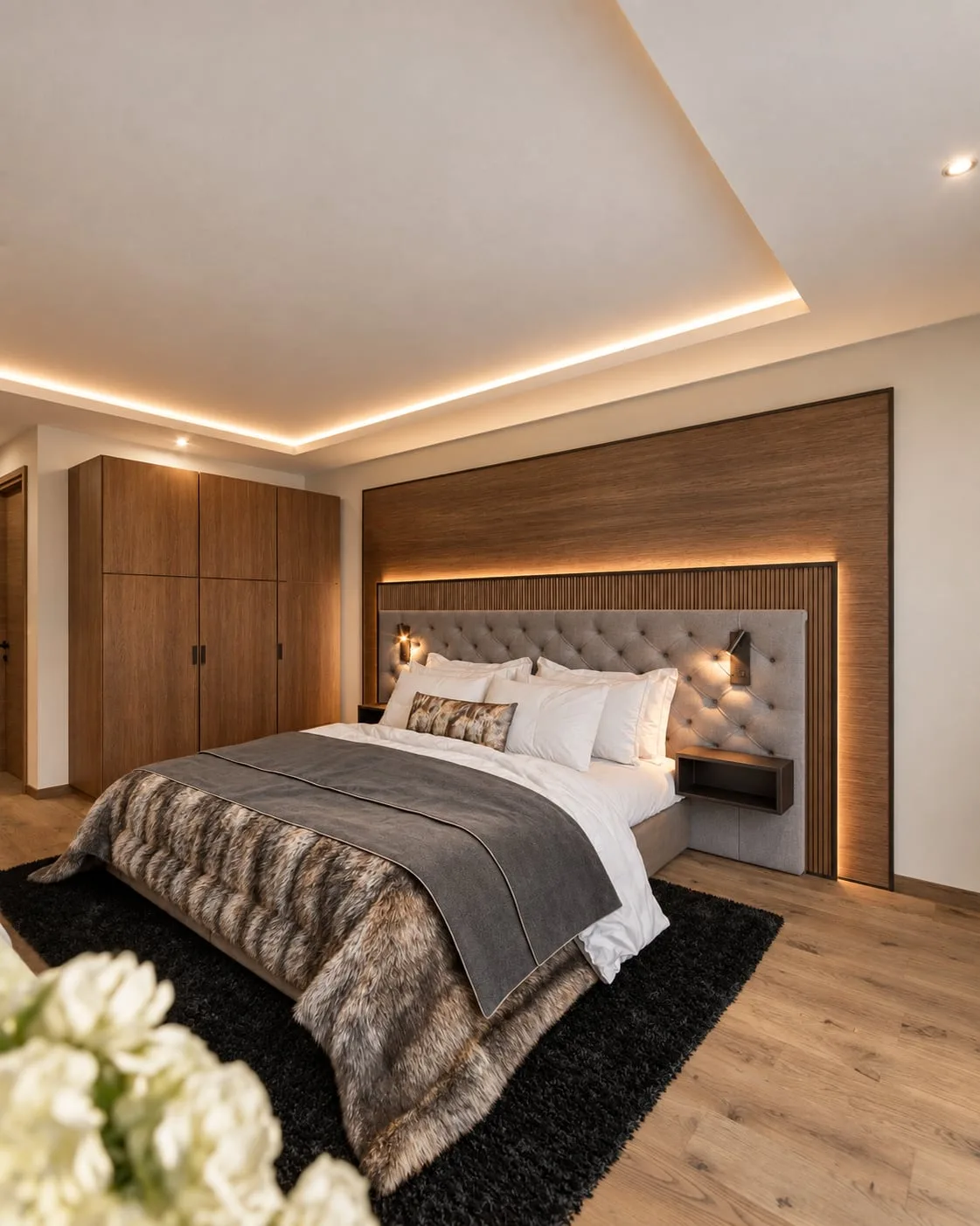 A bedroom in the upper apartment, fur throw across the bed, fitted wardrobe to one side and a backlit timber headboard wall