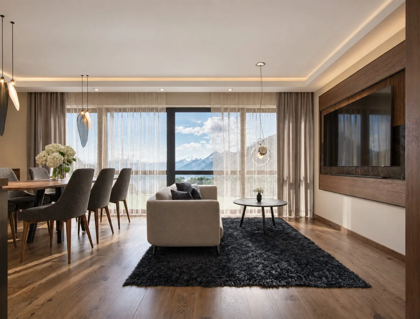 The living room from the kitchen end, dining table and chairs in front of a sofa on a dark rug, mountains through the glazing