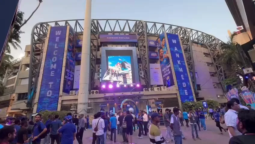 The front of Wankhede Stadium before a match, tall blue LED columns reading WELCOME TO and WANKHEDE either side of the main screen, crowd filling the forecourt